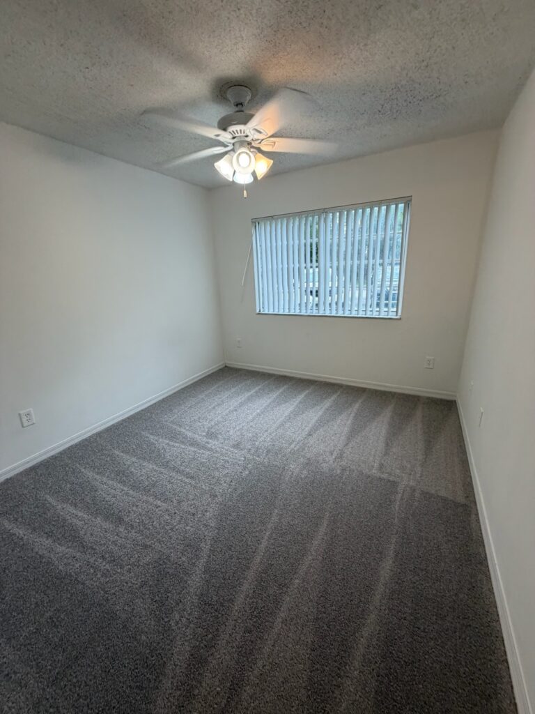 A bright, empty room in a home or apartment that has just been professionally cleaned. The space feels open, fresh, and full of possibility. Sunlight pours through sparkling clean windows, illuminating a gleaming floor with visible vacuum lines or a polished shine. The kitchen is visible through a doorway — counters bare and spotless, sink polished, and cabinets slightly ajar to show clean interiors. A single TFG Cleaning Services LLC branded door hanger hangs on the front door or entry closet, reading "Just Cleaned — Ready for Move-In." No boxes, no clutter, no furniture — just a pristine blank canvas waiting for its next chapter. The overall mood is one of cleanliness, anticipation, and the quiet satisfaction of a job done right.