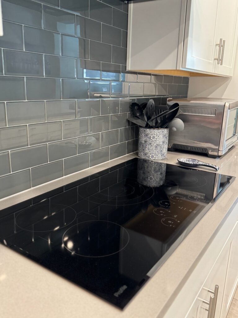 TFG CLEANING SERVICES LLC professional Cleaning in Sanford. Image: The image showcases a modern kitchen with a sleek black electric cooktop as the central feature. The cooktop is set on a light beige countertop, which contrasts with the dark gray subway tiles that form the backsplash behind it. A white microwave is positioned to the right of the cooktop, and a white utensil holder with black utensils is placed nearby. The kitchen cabinets are white, and the overall design is clean and minimalist.
